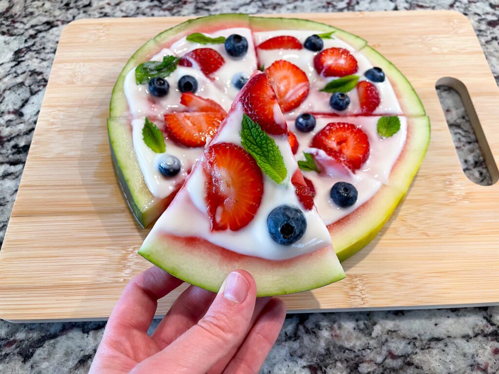 Holding a slice of dessert pizza with the rest of the dessert pizza behind it on the counter.